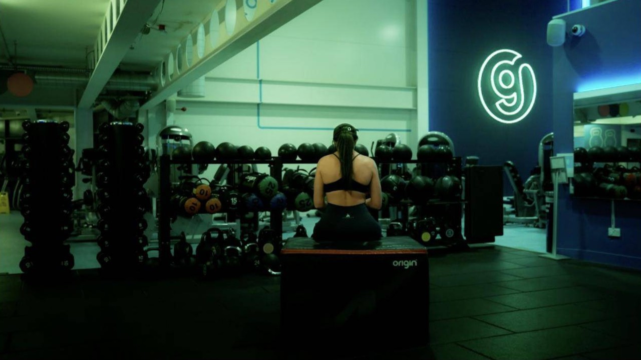 A woman sitting on a plyo box in a dimly lit gym, facing away from the camera